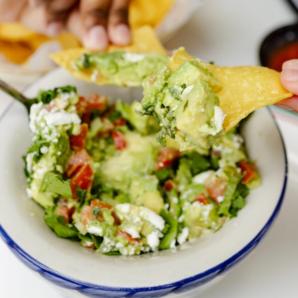 A hand dips a tortilla chip into a bowl of fresh guacamole with tomatoes and cheese.