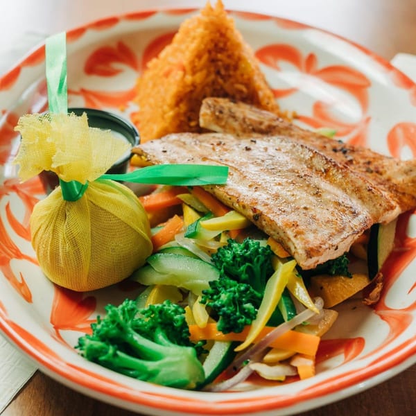 Grilled fish with rice, mixed vegetables, and a lemon wrapped in yellow cloth on a decorative plate.