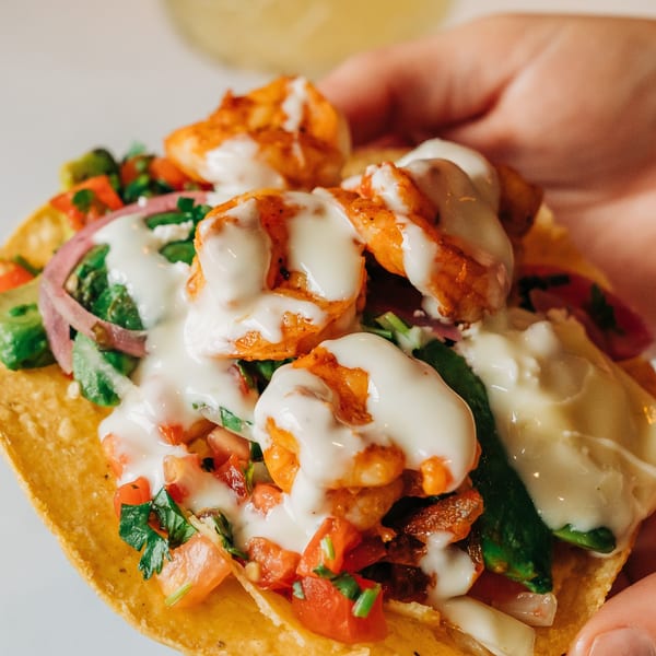 A hand holds a Si Señor Taqueria shrimp taco topped with white sauce, red onions, and salsa.